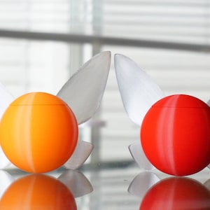 Puede incluir: Dos adornos decorativos en forma de huevo con alas. El adorno de la izquierda es naranja y el adorno de la derecha es rojo.