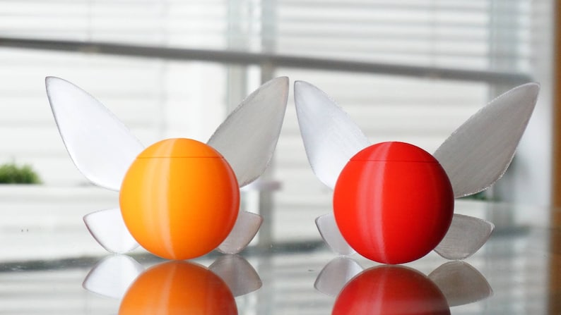 Puede incluir: Dos adornos decorativos en forma de huevo con alas. El adorno de la izquierda es naranja y el adorno de la derecha es rojo.