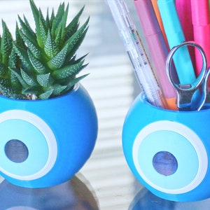 May include: Two blue ceramic planters with a white and light blue evil eye design. One planter has a small succulent plant inside, the other planter has a variety of colorful pens and a pair of silver scissors inside.
