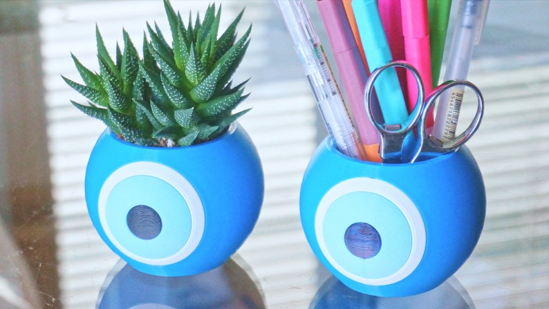 May include: Two blue ceramic planters with a white and light blue evil eye design. One planter has a small succulent plant inside, the other planter has a variety of colorful pens and a pair of silver scissors inside.