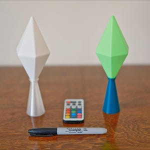 May include: Two geometric-shaped lamps, one white and one green and blue, stand on a wooden surface. A small remote control and a black Sharpie marker are between the lamps. The lamps have a diamond-like top and a conical base.