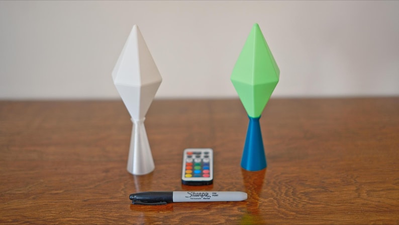 May include: Two geometric-shaped lamps, one white and one green and blue, stand on a wooden surface. A small remote control and a black Sharpie marker are between the lamps. The lamps have a diamond-like top and a conical base.
