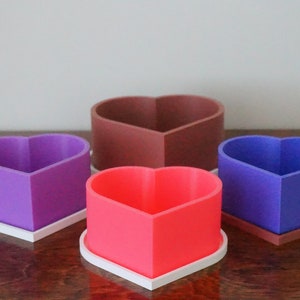 Puede incluir: Cuatro macetas en forma de coraz&oacute;n de diferentes colores: morado, marr&oacute;n, azul y rosa. Cada maceta tiene una base blanca.