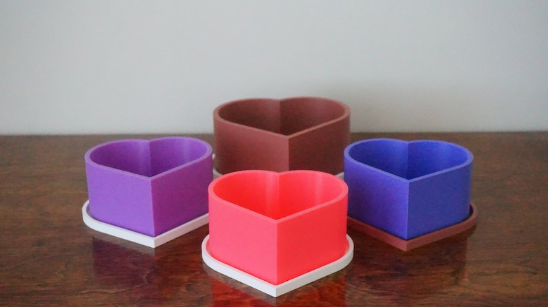 Puede incluir: Cuatro macetas en forma de coraz&oacute;n de diferentes colores: morado, marr&oacute;n, azul y rosa. Cada maceta tiene una base blanca.
