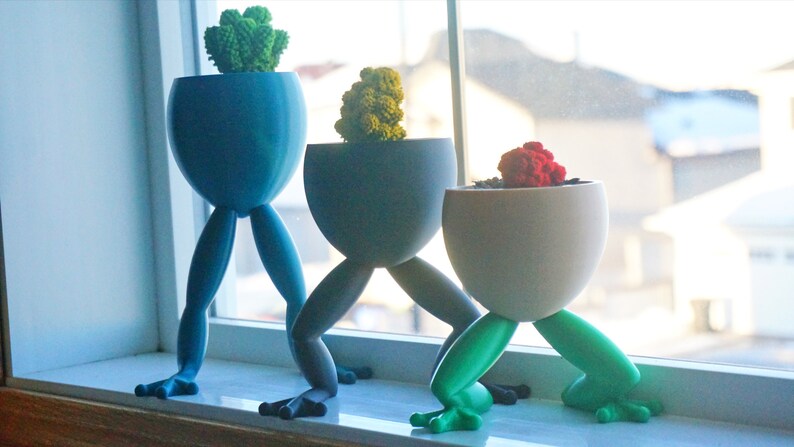 May include: Three frog-leg plant pots with different colored plants. The pots are blue, gray, and white. The plants are green, yellow-green, and red.