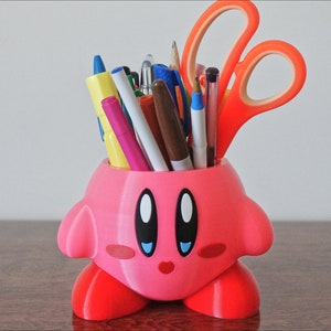 Puede incluir: Un porta l&aacute;pices rosa impreso en 3D con forma de personaje de dibujos animados con ojos grandes y una boca sonriente. El porta l&aacute;pices est&aacute; lleno de bol&iacute;grafos, l&aacute;pices y tijeras.