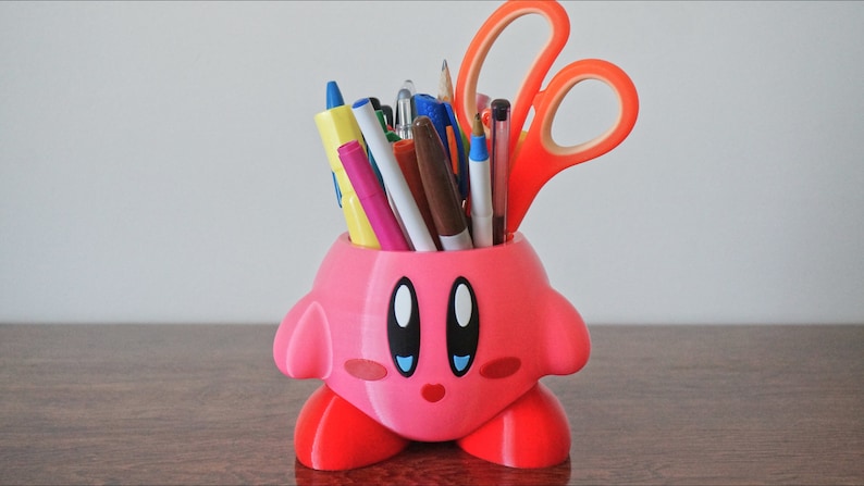 Puede incluir: Un porta l&aacute;pices rosa impreso en 3D con forma de personaje de dibujos animados con ojos grandes y una boca sonriente. El porta l&aacute;pices est&aacute; lleno de bol&iacute;grafos, l&aacute;pices y tijeras.