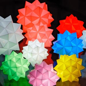 Pode incluir: Uma cole&ccedil;&atilde;o de oito lanternas de papel em forma de estrela geom&eacute;trica em v&aacute;rias cores, incluindo vermelho, branco, azul, verde, amarelo e rosa. As lanternas s&atilde;o iluminadas por dentro, projetando um brilho quente.