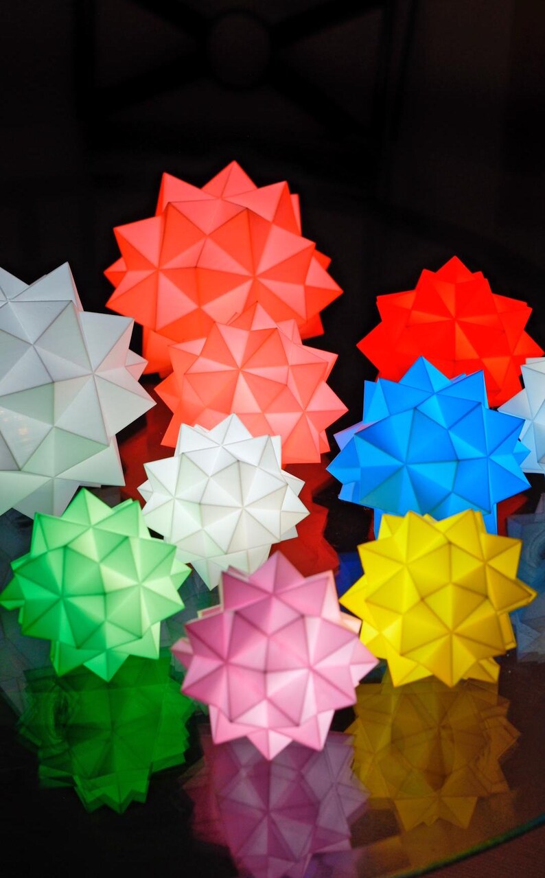 Pode incluir: Uma cole&ccedil;&atilde;o de oito lanternas de papel em forma de estrela geom&eacute;trica em v&aacute;rias cores, incluindo vermelho, branco, azul, verde, amarelo e rosa. As lanternas s&atilde;o iluminadas por dentro, projetando um brilho quente.