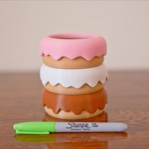 Puede incluir: Conjunto de tres macetas con forma de donut en rosa, blanco y marr&oacute;n con un marcador permanente Sharpie de punta fina verde.