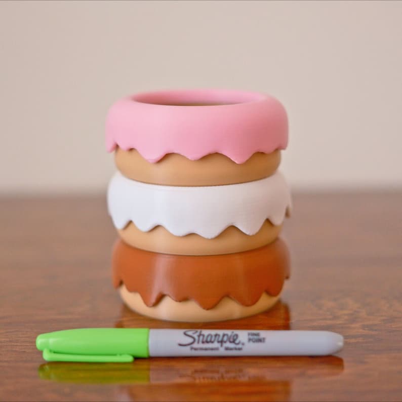 Puede incluir: Conjunto de tres macetas con forma de donut en rosa, blanco y marr&oacute;n con un marcador permanente Sharpie de punta fina verde.