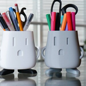 Op de afbeelding: Twee witte en grijze 3D-geprinte potloodhouders in de vorm van een cartoonfiguur met armen en benen. De houders hebben een zwart stopcontactontwerp aan de voorkant. De witte houder bevat een verscheidenheid aan pennen en potloden, terwijl de grijze houder kleurpotloden bevat.