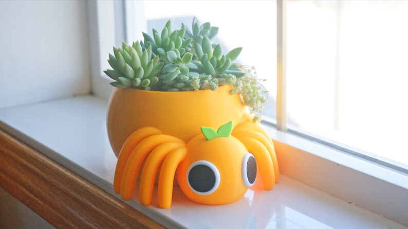 May include: A bright orange planter with a succulent plant sits on a windowsill next to an orange decorative spider. The spider has large black eyes and green leaf accents. The planter is round and has a smooth finish.