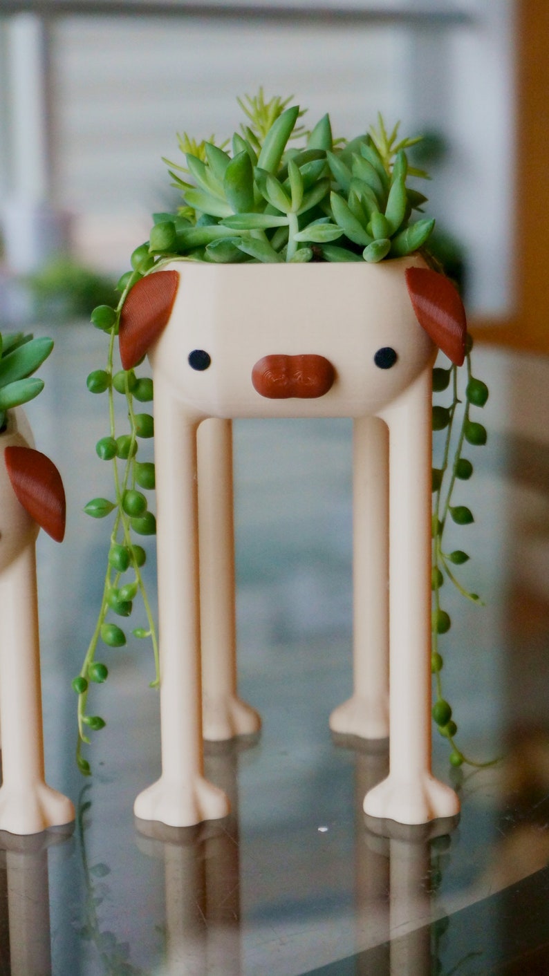 May include: A white ceramic planter shaped like a dog with brown ears and a nose. The planter has four legs and is holding a succulent plant with green leaves.
