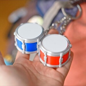 May include: Two miniature drum keychains. One drum is blue, the other is red, both with silver-colored hardware and white drumheads. The keychains are held in a hand, with a blurred background.