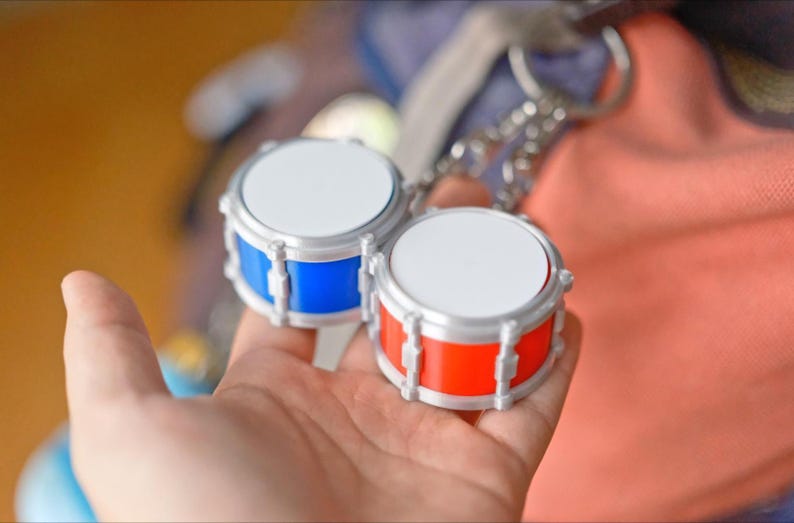 May include: Two miniature drum keychains. One drum is blue, the other is red, both with silver-colored hardware and white drumheads. The keychains are held in a hand, with a blurred background.