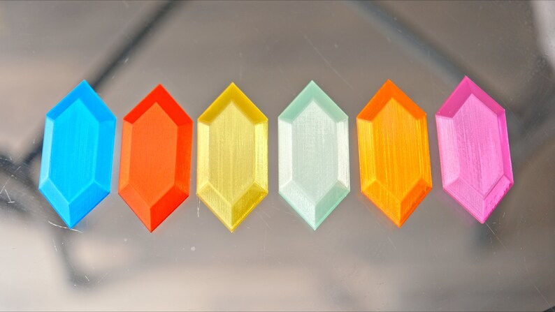 May include: Six different colored plastic gems in the shape of a hexagon. The gems are arranged in a row from left to right: blue, orange, yellow, light green, orange, and pink.