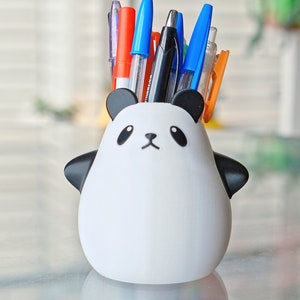 Op de afbeelding: Een witte keramische panda-vormige bureau-organizer met zwarte oren, ogen en neus. De organizer is gevuld met een verscheidenheid aan pennen en potloden, waaronder blauw, rood en oranje.