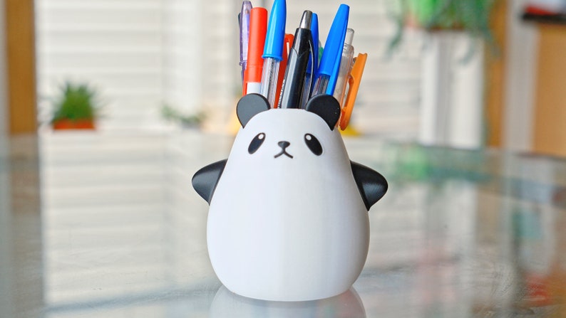 Op de afbeelding: Een witte keramische panda-vormige bureau-organizer met zwarte oren, ogen en neus. De organizer is gevuld met een verscheidenheid aan pennen en potloden, waaronder blauw, rood en oranje.