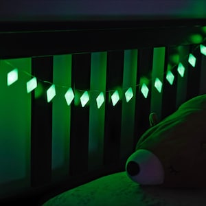May include: A string of glowing green diamond-shaped lights hangs over a bed. The lights are strung across a dark wooden headboard. A large, plush, brown bear-shaped pillow rests on the bed. The scene is dimly lit with a green glow.