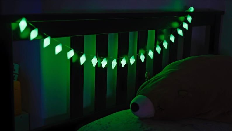 May include: A string of glowing green diamond-shaped lights hangs over a bed. The lights are strung across a dark wooden headboard. A large, plush, brown bear-shaped pillow rests on the bed. The scene is dimly lit with a green glow.