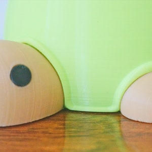 May include: A green and brown turtle-shaped planter with a black eye. The planter has a smooth, rounded surface and a textured base.