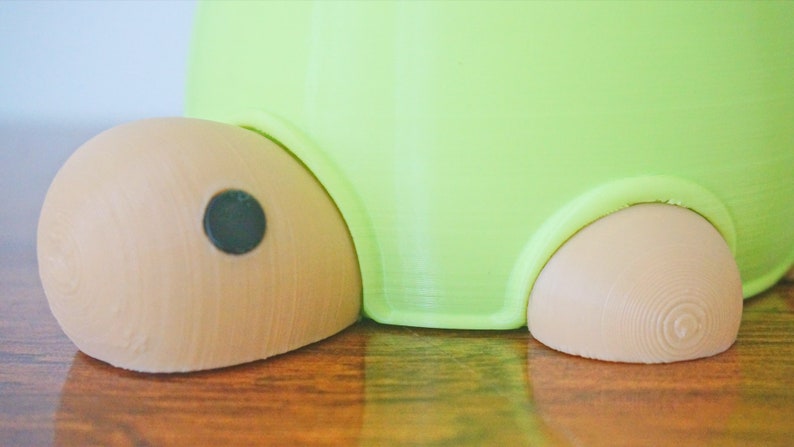 May include: A green and brown turtle-shaped planter with a black eye. The planter has a smooth, rounded surface and a textured base.