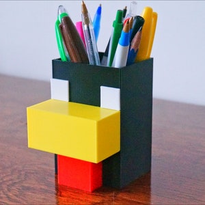 May include: A black, yellow, and red 3D printed pen holder shaped like a Minecraft character. The holder has a square base and is filled with pens and pencils.