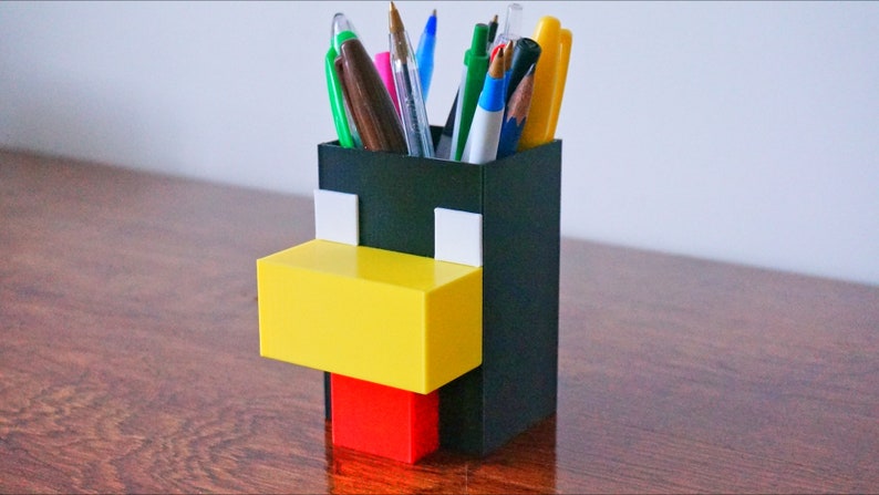 May include: A black, yellow, and red 3D printed pen holder shaped like a Minecraft character. The holder has a square base and is filled with pens and pencils.