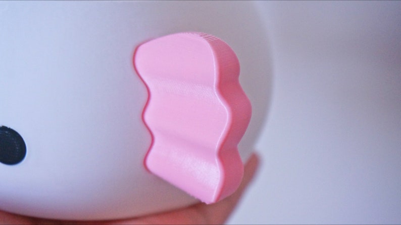 K&ouml;nnte beinhalten: Eine wei&szlig;e Keramiktasse mit einem rosa, wellenf&ouml;rmigen, 3D-Ohr. Die Tasse hat ein schwarzes, kreisf&ouml;rmiges Auge.