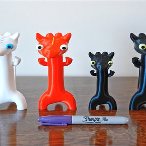 May include: Four colorful, stylized dragon figurines in white, red, black, and black with a purple Sharpie marker for scale. The dragons have large, expressive eyes and a unique, elongated body shape. They stand on a wooden surface.