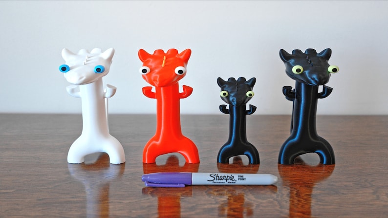 May include: Four colorful, stylized dragon figurines in white, red, black, and black with a purple Sharpie marker for scale. The dragons have large, expressive eyes and a unique, elongated body shape. They stand on a wooden surface.