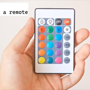 May include: A white remote control with colored buttons is held in a hand. The remote has buttons labeled "OFF" and "ON", and also includes color options and special effects like "FLASH" and "FADE". The text "RC version Comes with a remote" is visible.