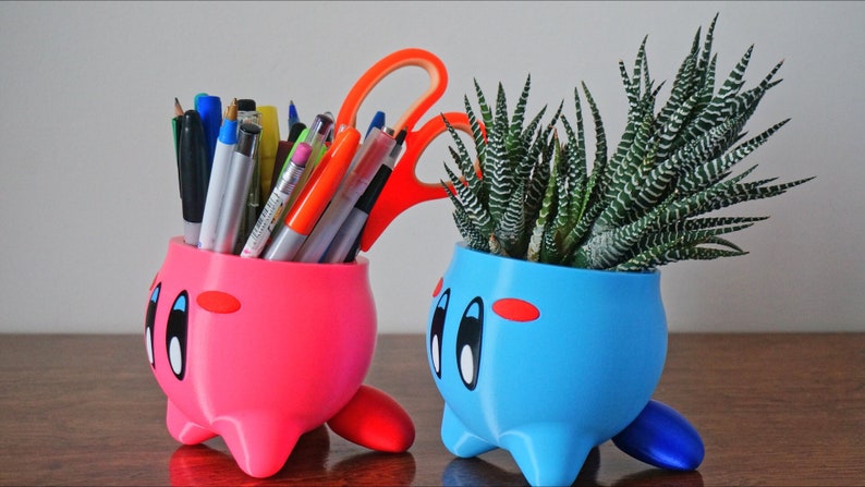 May include: Two 3D printed character planters, one pink and one blue, with a cartoon face. The pink planter is filled with pens and pencils, while the blue planter holds a green succulent plant.