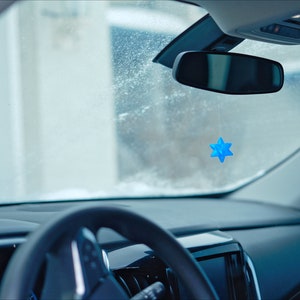 May include: A blue star-shaped car air freshener hanging from the rearview mirror of a car. The windshield is covered in frost.