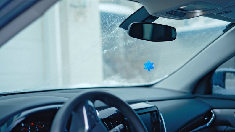 May include: A blue star-shaped car air freshener hanging from the rearview mirror of a car. The windshield is covered in frost.