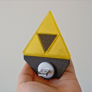May include: A yellow plastic triangular-shaped USB drive with a black inner triangle and a white USB connector. The drive is shaped like the Triforce from the Legend of Zelda video game series.