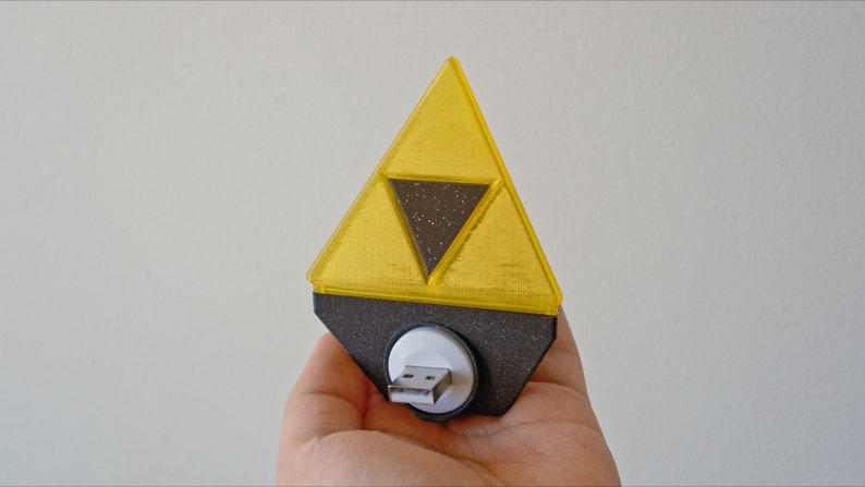 May include: A yellow plastic triangular-shaped USB drive with a black inner triangle and a white USB connector. The drive is shaped like the Triforce from the Legend of Zelda video game series.