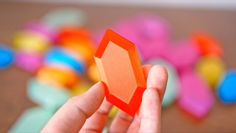 May include: A bright orange, geometrically shaped, plastic gem. The gem is held in a hand, with a blurred background of colorful, geometric shapes.