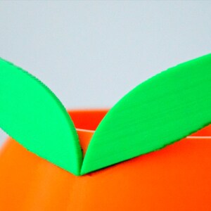 May include: A bright orange ceramic planter with two green felt leaves attached to the top.