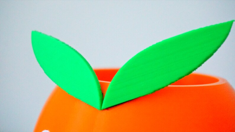 May include: A bright orange ceramic planter with two green felt leaves attached to the top.
