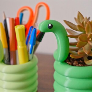 May include: Two green, snake-shaped desk organizers. One holds pens, pencils, and scissors. The other contains a succulent plant. The organizers are a bright green color and have a coiled design. The snake heads have a black and white eye.