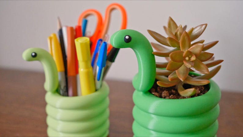 May include: Two green, snake-shaped desk organizers. One holds pens, pencils, and scissors. The other contains a succulent plant. The organizers are a bright green color and have a coiled design. The snake heads have a black and white eye.