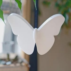 May include: A white butterfly-shaped decoration hangs in front of a blurred background. The butterfly has a symmetrical design with rounded wings. Green leaves are visible in the foreground and background, suggesting a nature-inspired theme.