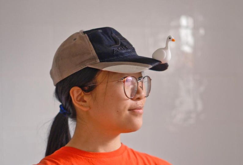 May include: A baseball cap with a tan and navy blue design, featuring a small white goose figurine perched on the brim. The cap is worn by a person wearing glasses and an orange shirt.