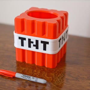 K&ouml;nnte beinhalten: Ein roter und wei&szlig;er Plastikbeh&auml;lter in Form eines Minecraft TNT-Blocks. Der Beh&auml;lter hat ein wei&szlig;es Band um sich herum mit dem Wort "THT" in schwarzen Buchstaben.