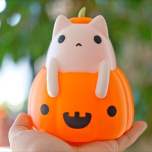 May include: A Halloween-themed decorative light featuring a white cat figure perched inside an orange pumpkin. The pumpkin has a carved jack-o'-lantern face with black eyes and a smiling mouth. The cat has black eyes and is holding onto the pumpkin.