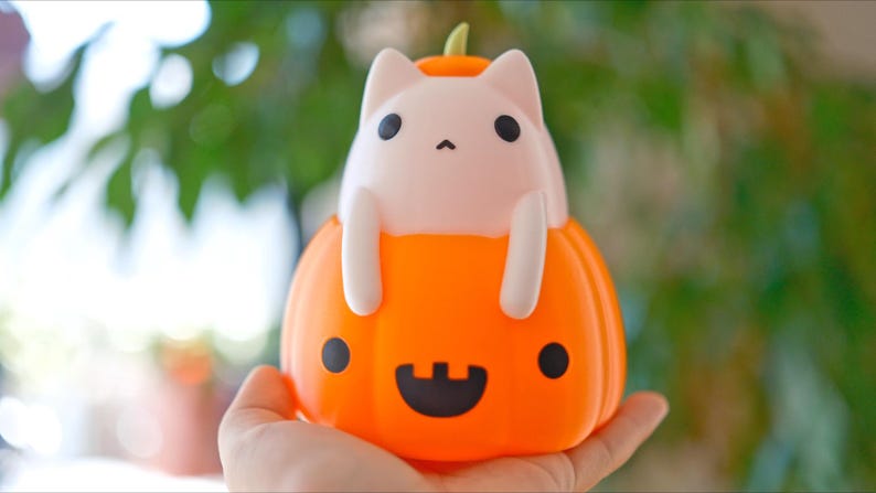 May include: A Halloween-themed decorative light featuring a white cat figure perched inside an orange pumpkin. The pumpkin has a carved jack-o'-lantern face with black eyes and a smiling mouth. The cat has black eyes and is holding onto the pumpkin.
