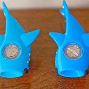 May include: Two blue whale shark shaped toys with a white spot on each side. The toys have a clear plastic circle in the center with a silver screw in the middle.