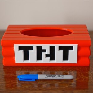 May include: A red plastic container with a round opening and the word "THT" printed on the front. The container is sitting on a wooden surface.